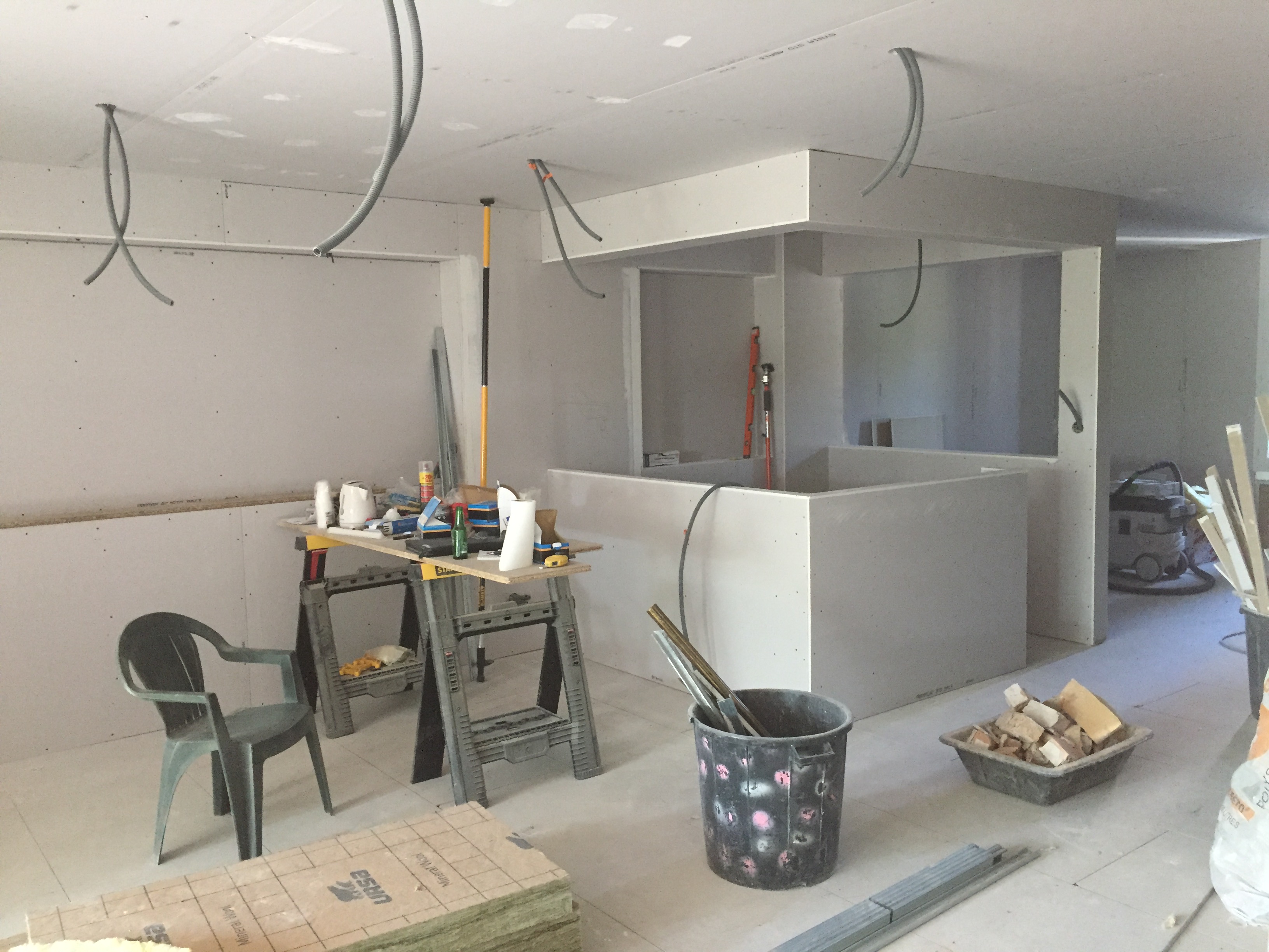 Chantier placo : Pose des cloisons sèches et îlot de cuisine. Intérieur de chantier cuisine/bar : cloisons sèches posées, gaines électriques, isolant URSA. Outils et seau sur sol carrelé.