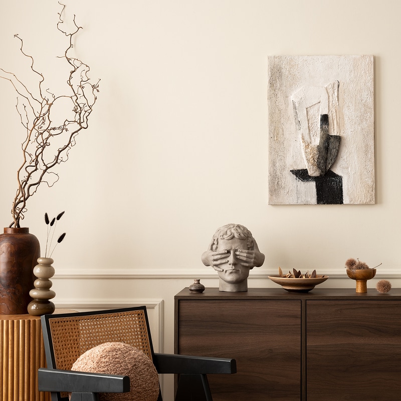 Décoration moderne minimaliste : mur beige, cabinet foncé, fauteuil en rotin, tête sculptée (mains sur les yeux), et tableau texturé.