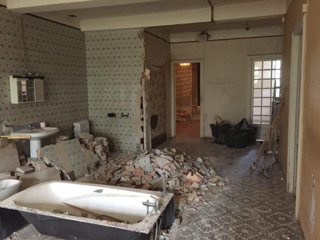Démolition de salle de bain : baignoire, lavabo, et un grand tas de gravats sur sol carrelé. Vieux papier peint à motifs et murs exposés.