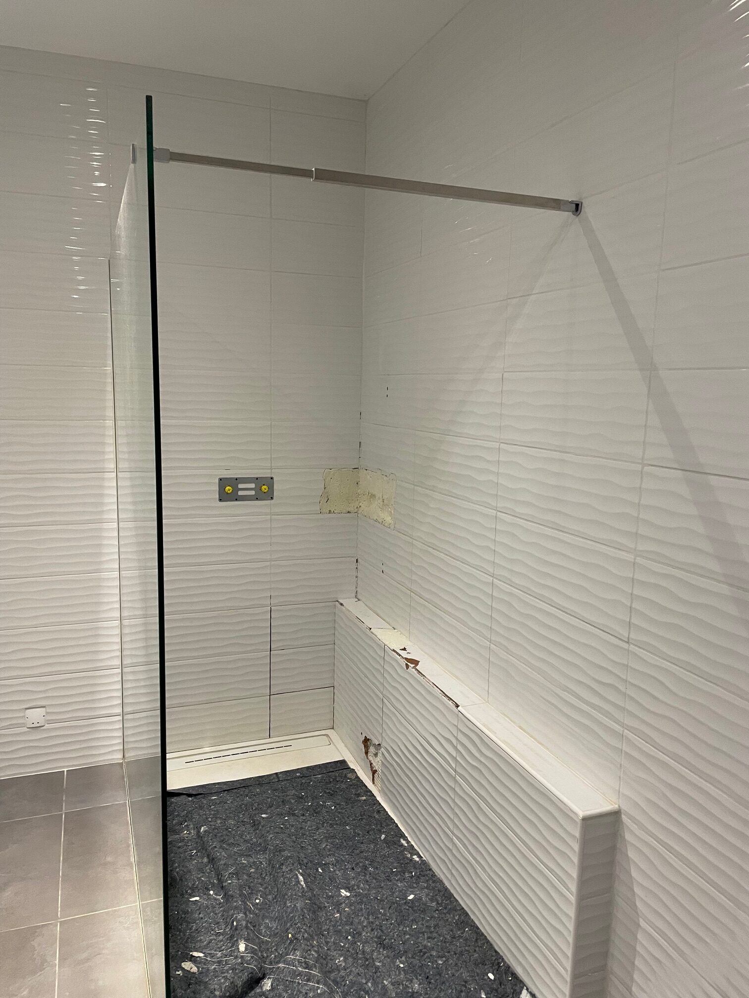 Douche italienne : carrelage blanc texturé vague, travaux Douche italienne moderne en construction. Carrelage mural blanc à effet vague 3D, pare-douche en verre et support de robinetterie fixé.