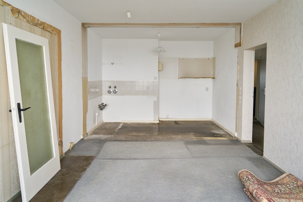 Pièce vide en démolition. Moquette grise soulevée, porte intérieure blanche déposée. L'embrasure mène à l'ancienne cuisine carrelée.