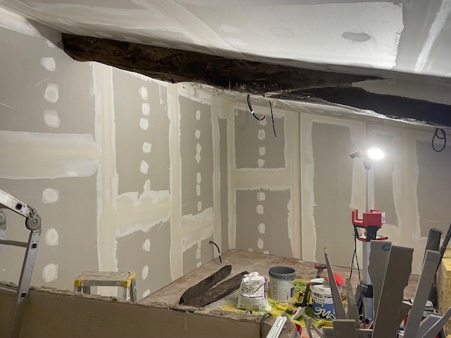 Rénovation Placo : Installation et finition des murs intérieurs. Chantier intérieur : plaques de plâtre posées et enduites, révélant une vieille poutre en bois sombre. Éclairage de travail et outils de finition.