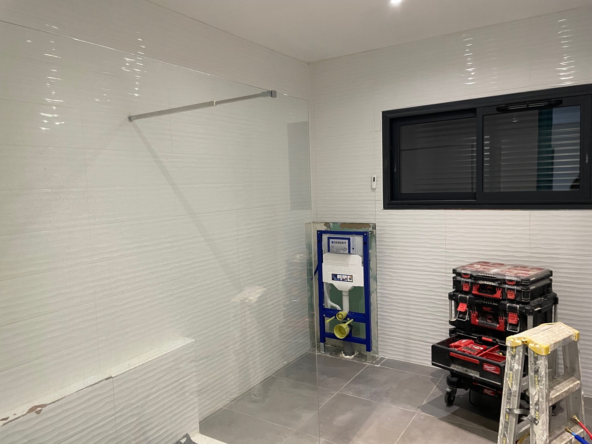 Rénovation salle de bain : Geberit, carrelage texturé et fenêtre noire. Rénovation salle de bain. Carrelage blanc texturé, sol gris, cloison de verre. Installation du châssis WC GEBERIT. Outils Milwaukee.
