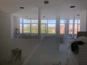 Intérieur d'un bâtiment en chantier avec murs blancs, grandes fenêtres et matériel de construction.