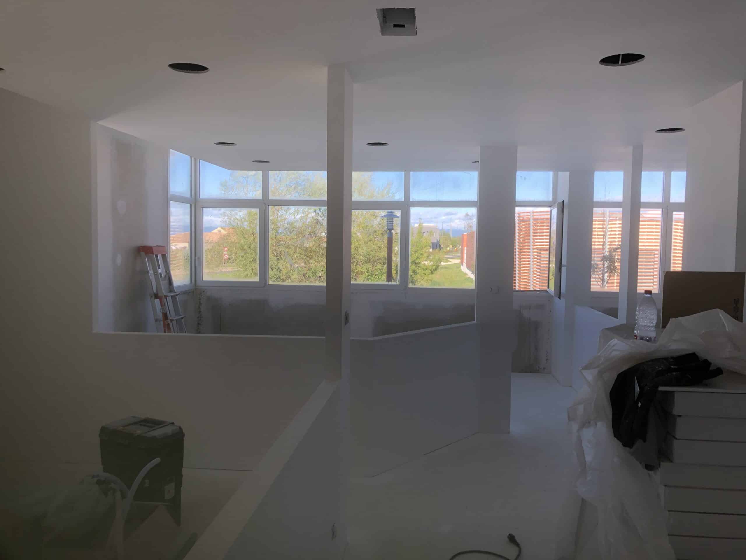 Intérieur d'un bâtiment en chantier avec murs blancs, grandes fenêtres et matériel de construction.
