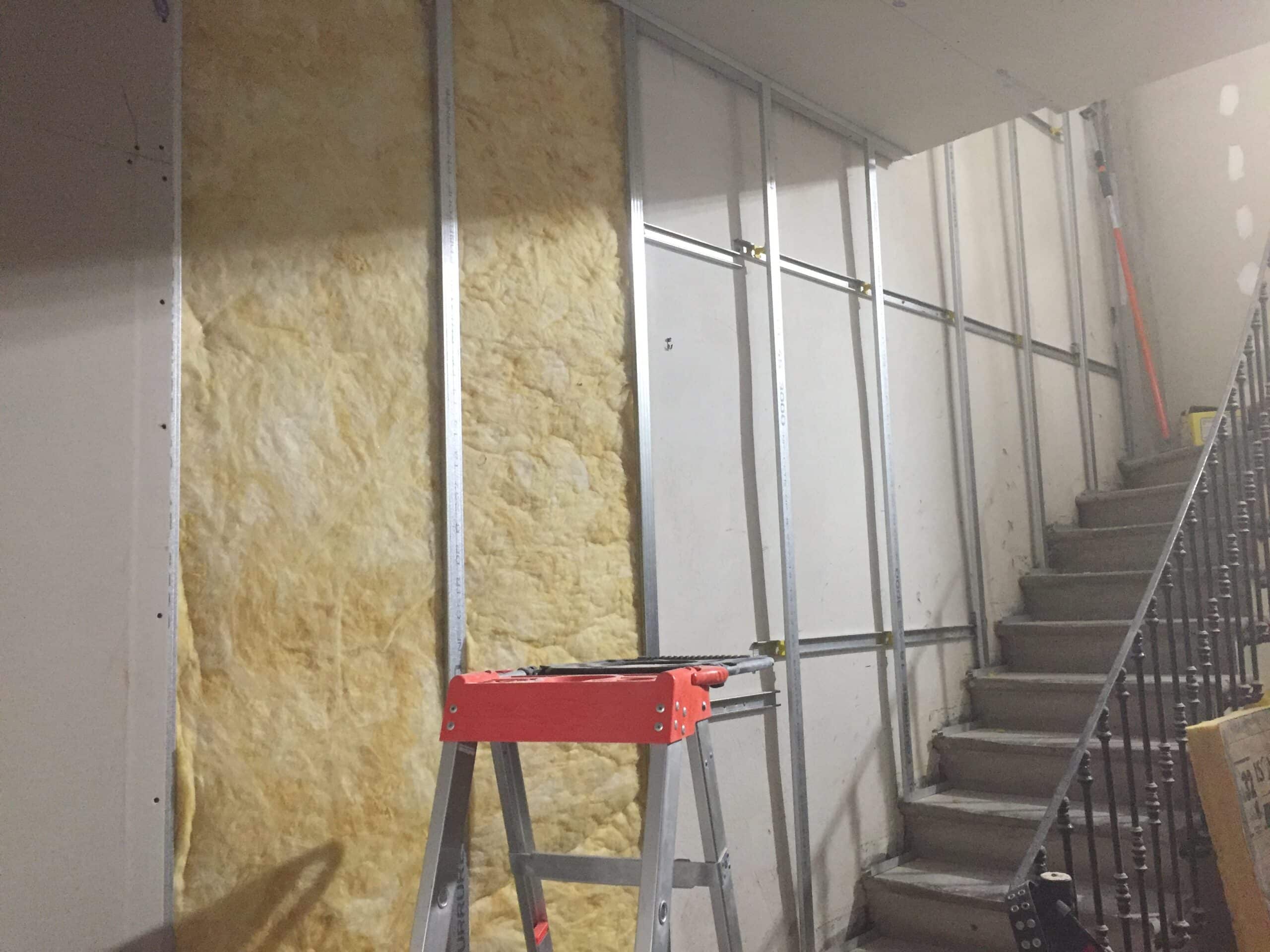 Pose de laine de verre et rails métalliques pour l'isolation d'un mur intérieur à côté d'un escalier en chantier.