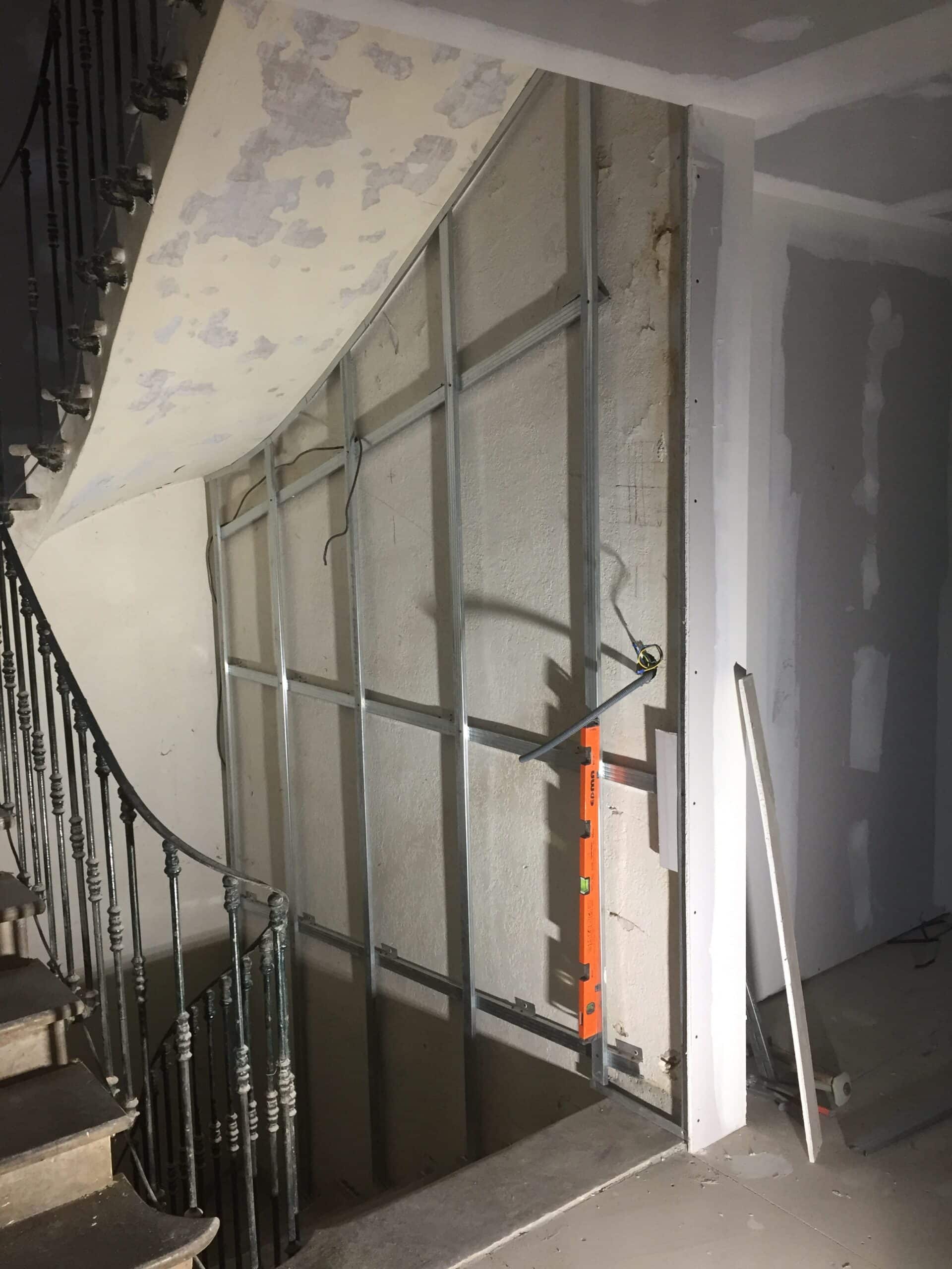 Ossature métallique pour cloison sèche sous un escalier en rénovation avec niveau à bulle et câbles électriques.