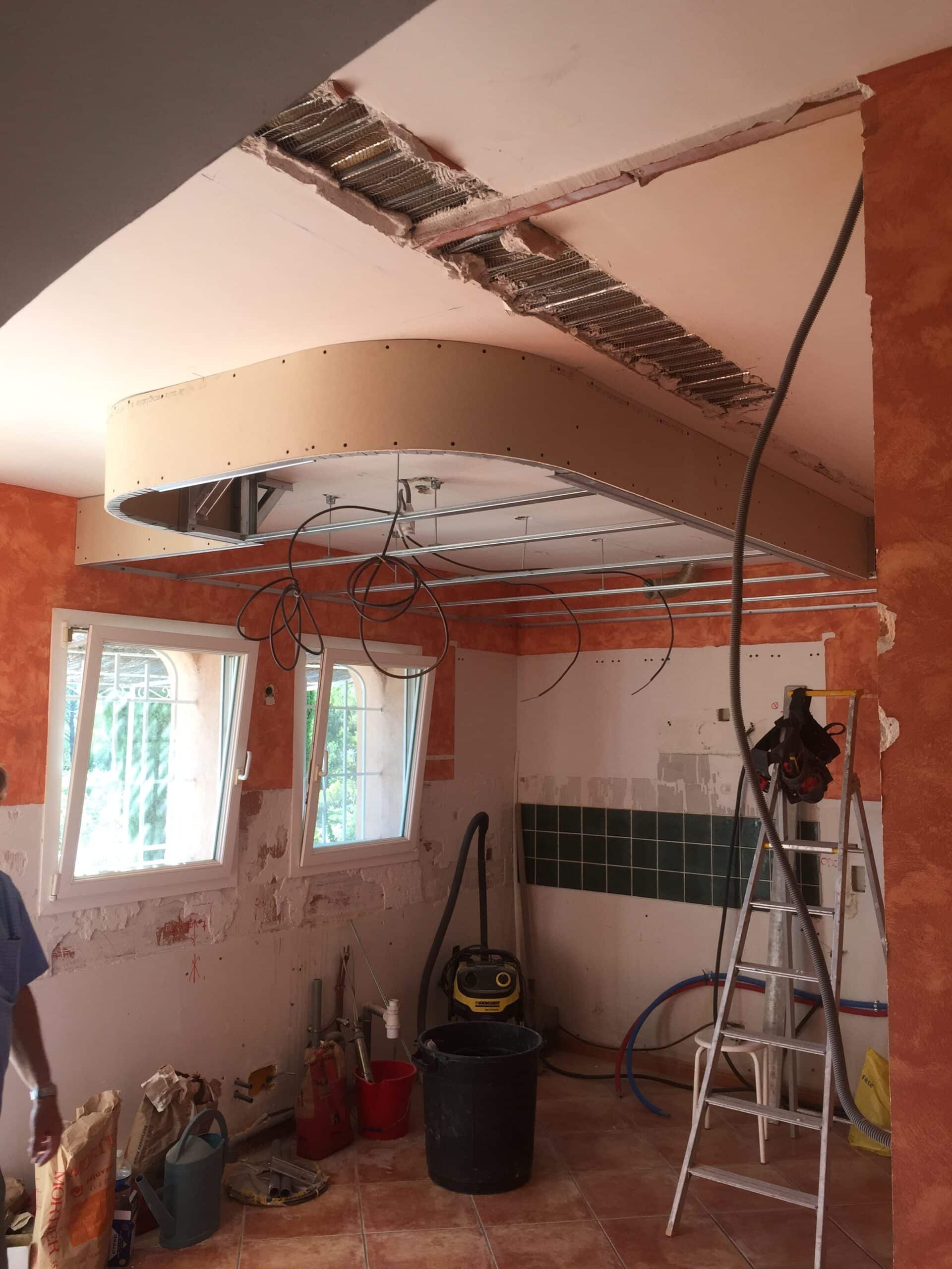 Chantier de rénovation avec faux plafond arrondi en placo, câbles électriques, échelle et outils dans une cuisine.