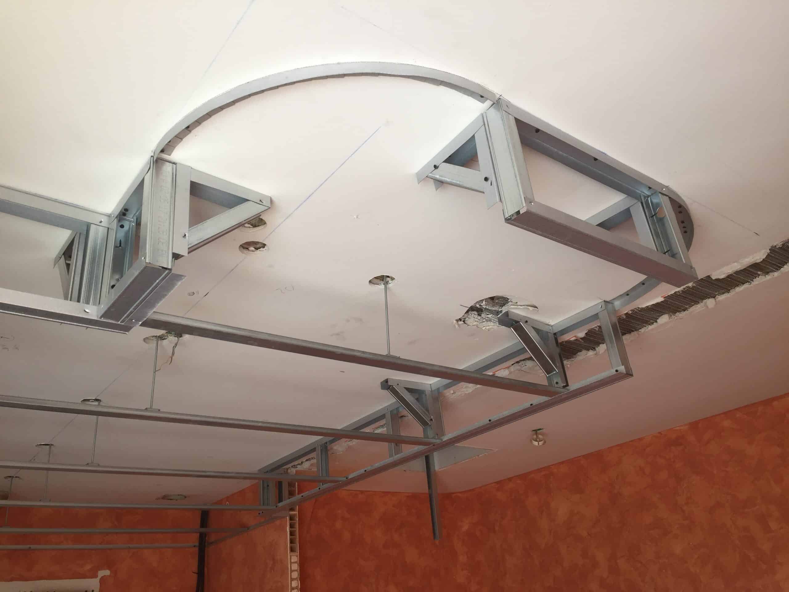 Structure métallique de faux plafond suspendu avec rails en métal et courbes sur plafond blanc et mur orange.